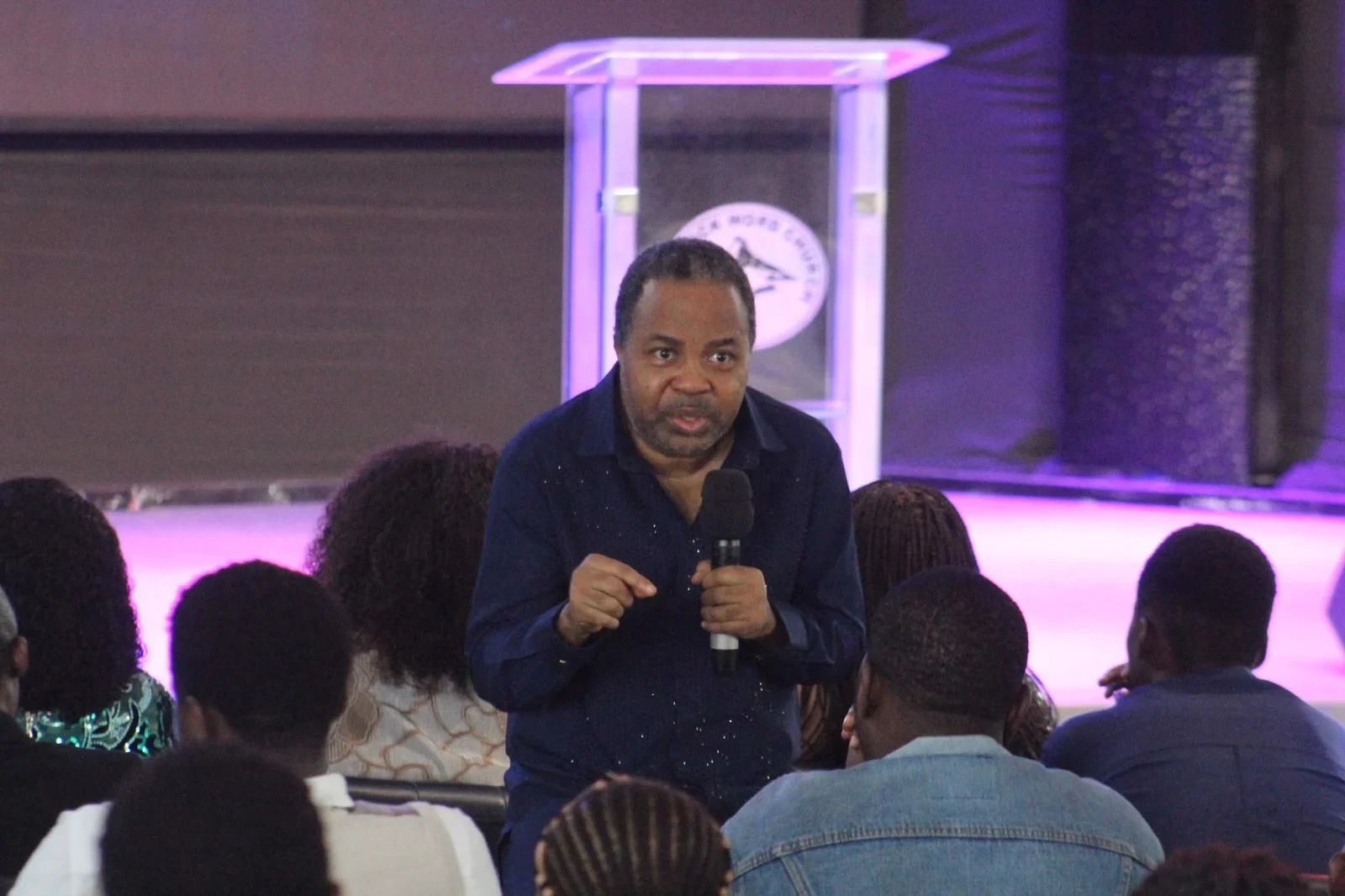 Reverend Aromaegbe leaning towards seated listeners while speaking into a microphone.