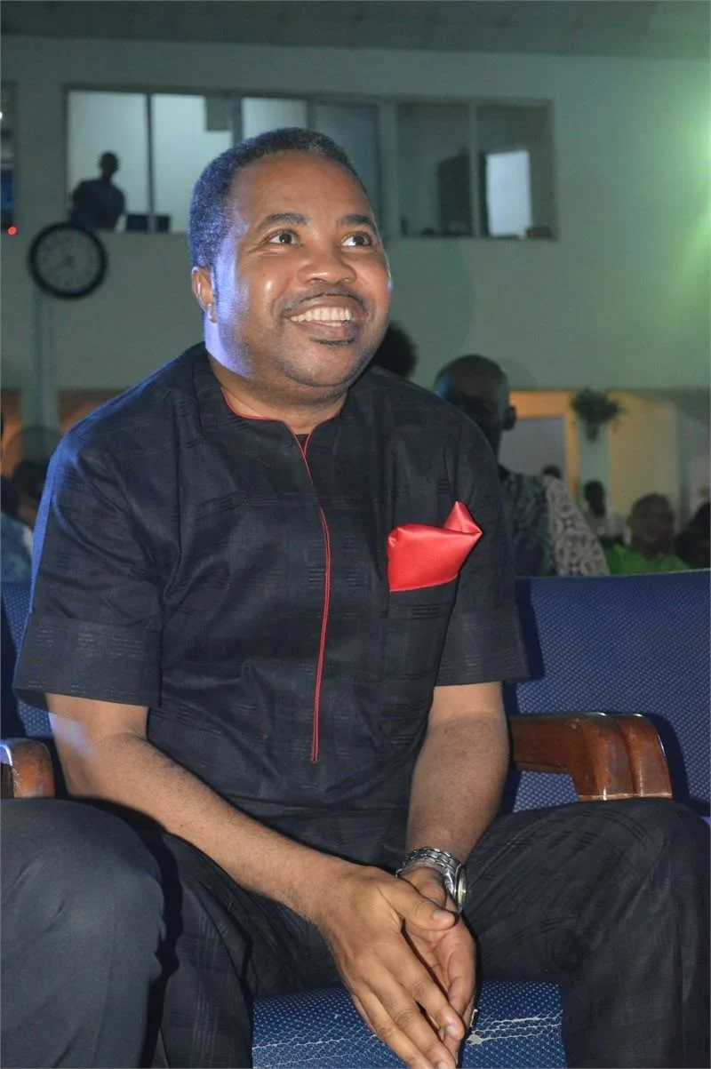 Reverend Aromaegbe seated in a black outfit with a red pocket square, smiling and looking upwards.