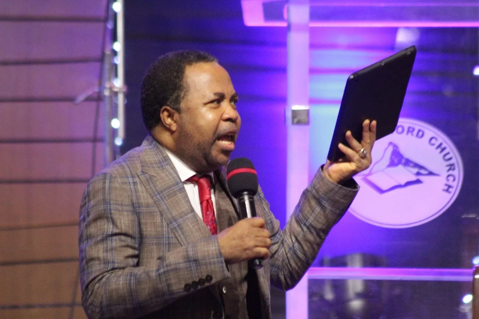 Reverend Aromaegbe speaking into a microphone, holding up a tablet, in a checked jacket and red tie.